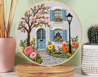 May include: A detailed embroidery hoop art piece featuring a white house with blue shutters and door, a flowering tree, and a stone path. The scene is filled with colorful flowers in pink, yellow, and orange. A black lamppost stands to the right.