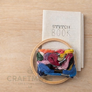 Puede incluir: Un kit de manualidades con un bastidor de bordado de madera, una pila de páginas en blanco con las palabras "STITCH BOOK" en negro y una bolsa de plástico transparente llena de hilo de bordar colorido. El kit está sobre una superficie de madera clara.