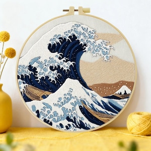 Op de afbeelding: Geborduurd kunstwerk van Hokusai's "De Grote Golf van Kanagawa" in een ronde houten hoepel. Het ontwerp toont een grote, donkerblauwe golf met witte kammen, een boot en een berg op de achtergrond. De borduursel is op witte stof.