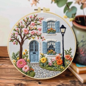 以下が含まれることがあります： 愛らしいコテージのシーンを描いた刺繍フープアート。青いドアとシャッター、花の咲く木、石畳の小道がデザインに含まれています。木製のフープに額装されており、さまざまなカラフルな花が特徴です。