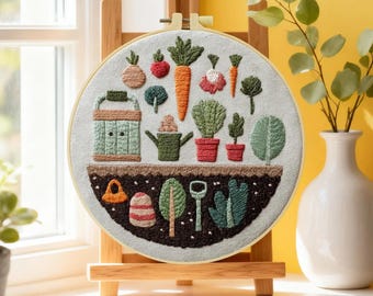 May include: Embroidered hoop art featuring a garden scene with vegetables, plants, and gardening tools. The design includes a carrot, tomato, watering can, and other garden elements. The artwork is displayed on a wooden easel.