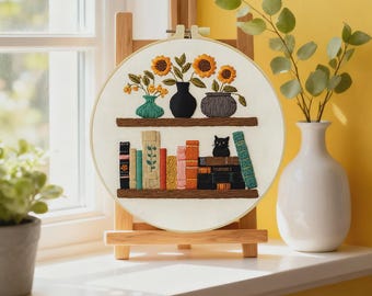May include: Embroidered hoop art depicting a bookshelf. The top shelf has three vases with sunflowers and other flowers. The bottom shelf shows a collection of books and a small black cat. The artwork is displayed on a wooden easel.