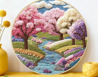 May include: A detailed, hand-stitched embroidery hoop art piece. The artwork depicts a vibrant landscape with a river, colorful trees, and flowers. The color palette includes pink, green, blue, and purple. The embroidery is set in a wooden hoop.