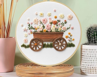 Pode incluir: Arte de bordado floral em bastidor, apresentando uma carroça de madeira cheia de flores coloridas. A carroça tem rodas marrons e está sobre um fundo branco. As flores são em tons de rosa, amarelo e laranja, com folhas verdes.
