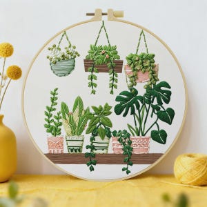 Pode incluir: Arte de bordado em bastidor com uma coleção de plantas verdes em vários vasos e jardineiras suspensas. As plantas incluem trepadeiras e variedades de folhas grandes. A obra de arte é definida contra um fundo branco, com uma prateleira de madeira e um bastidor de madeira natural.