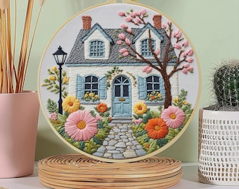 Pode incluir: Cena de casa bordada em um círculo. A casa tem um telhado cinza, persianas azuis e uma porta azul. Flores rosa, laranja e amarelas cercam um caminho de pedra que leva à casa. Uma árvore com flores rosa está à direita.