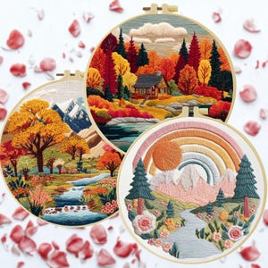 May include: Three circular embroidery hoops featuring landscape scenes. One shows a cabin in a forest with autumn colours. Another depicts a mountain scene with a river and trees. The third features a rainbow over a mountain range.