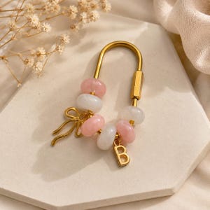 Può includere: Un portachiavi dorato a forma di U con perline rotonde alternate rosa e bianche. Il portachiavi include un ciondolo a forma di fiocco dorato e un ciondolo a forma di lettera B appeso alla base. Chiusura con vite cilindrica.