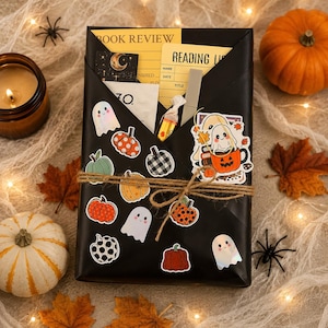 May include: A black envelope-style package decorated with stickers of ghosts and pumpkins, tied with twine. Inside are a yellow book review card, a reading list card, a small candy, and a ghost sticker. The package is surrounded by autumn leaves.