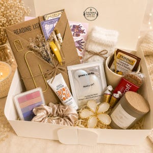 May include: A gift box containing a brown envelope labeled Reading List, a pair of white fuzzy socks, a mug with Twinings tea, a Biscoff cookie, a shea butter hand cream, Grace & Stella eye masks, a pink and purple palette, a scrunchie, and a candle.