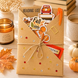May include: A brown kraft paper envelope labeled READING LIST, decorated with small colorful rhinestones and tied with twine. It contains autumn-themed stickers, a small orange pen, and a Twinings tea bag, creating a cozy seasonal stationery set.