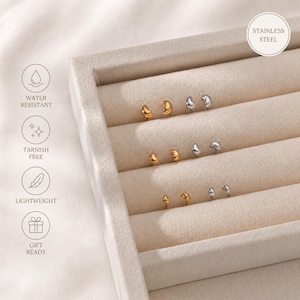 May include: A display of small stud earrings in gold and silver tones, arranged in a beige jewelry tray. The earrings have a smooth, rounded design. The image includes icons indicating water resistance, tarnish-free, lightweight, and gift-ready features.