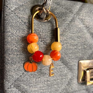 May include: A gold-toned metal carabiner clip attached to a gray quilted fabric surface. The clip features a series of round beads in shades of orange, yellow, and red, along with a small orange pumpkin charm and a gold-toned letter E charm.