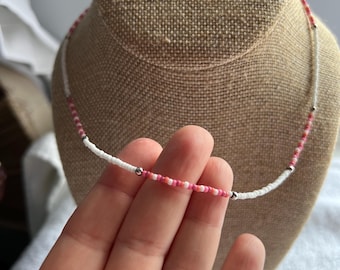 May include: A delicate beaded necklace with a mix of white, pink, and red beads. The necklace is displayed on a neutral-toned bust, showcasing its design. The beads are small and evenly spaced, creating a subtle, elegant accessory.