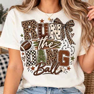 May include: A cream-colored t-shirt featuring the text "RUN the DANG Ball" in a mix of leopard print, checkered, and solid brown patterns. The design includes a football graphic, a large leopard print bow, small stars, and hearts.