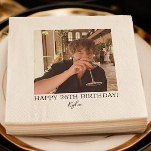 May include: A stack of white paper napkins featuring a photo of a person with a glass of wine. The text "HAPPY 26TH BIRTHAY! Kyle" is printed below the photo. The napkins are on a plate with a gold rim, set for a celebration.