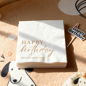 Peut inclure: Une pile de serviettes en papier blanches avec l'inscription "HAPPY birthday" en lettres dor&eacute;es. Le nom "BROCK" et la date "04.06 2026" sont &eacute;galement imprim&eacute;s. Un petit cake topper "Happy Birthday" est visible.
