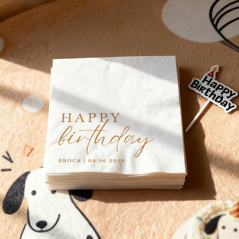 Peut inclure: Une pile de serviettes en papier blanches avec l'inscription "HAPPY birthday" en lettres dor&eacute;es. Le nom "BROCK" et la date "04.06 2026" sont &eacute;galement imprim&eacute;s. Un petit cake topper "Happy Birthday" est visible.