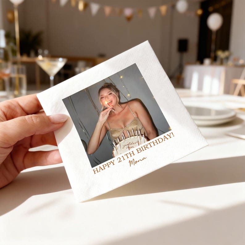 May include: A white square napkin with a printed photo of a woman in a gold dress holding a lit sparkler. The text "HAPPY 21TH BIRTHDAY! Mona" is printed in gold. The napkin is held by a person.