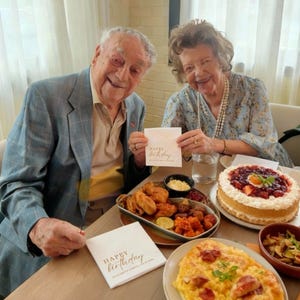 Peut inclure: Une c&eacute;l&eacute;bration d'anniversaire avec un couple tenant des serviettes avec l'inscription "Happy Birthday". La table est garnie d'un g&acirc;teau aux fraises, de poulet frit et de divers autres plats. L'homme porte un blazer bleu.