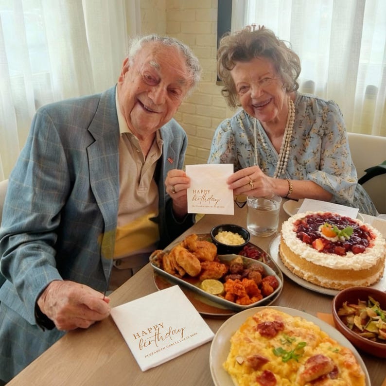 Peut inclure: Une c&eacute;l&eacute;bration d'anniversaire avec un couple tenant des serviettes avec l'inscription "Happy Birthday". La table est garnie d'un g&acirc;teau aux fraises, de poulet frit et de divers autres plats. L'homme porte un blazer bleu.