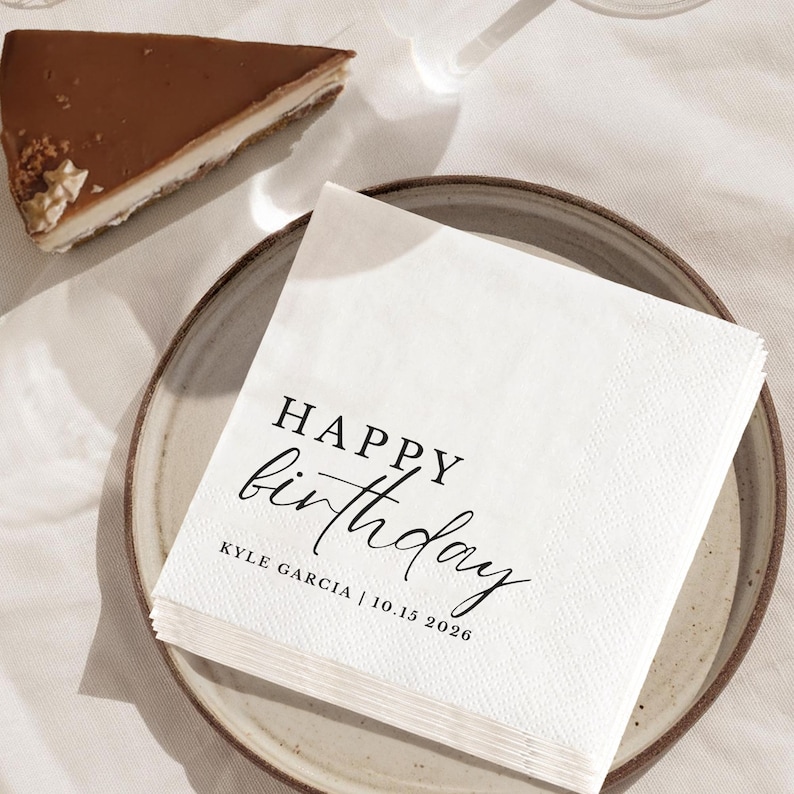 Peut inclure: Une pile de serviettes en papier blanches avec l'inscription "HAPPY birthday" en &eacute;criture noire, ainsi que le nom "KYLE GARCIA | 10.15.2026". Les serviettes sont pos&eacute;es sur une assiette en c&eacute;ramique, avec une part de g&acirc;teau au chocolat en arri&egrave;re-plan.