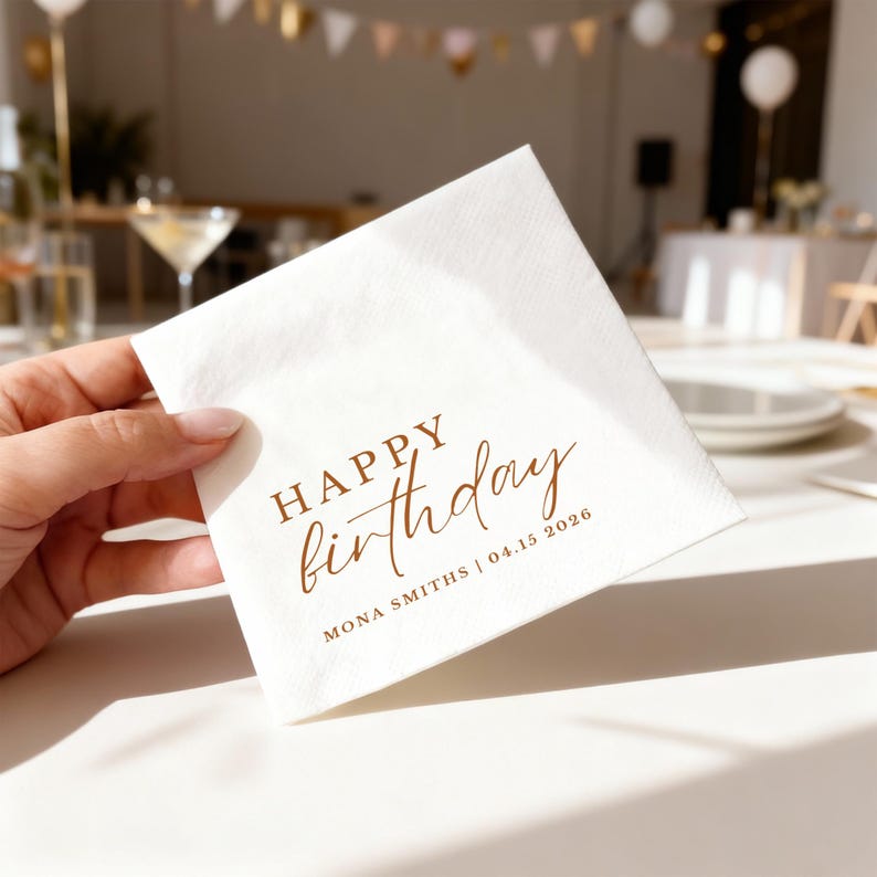 Peut inclure: Une serviette carr&eacute;e blanche avec les mots "HAPPY birthday" imprim&eacute;s en marron. En dessous, on peut lire "MONA SMITHS | 04.15 2026". La serviette est tenue par une main, avec un arri&egrave;re-plan flou d'une f&ecirc;te.