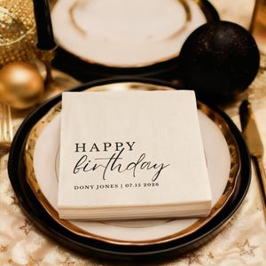 Peut inclure: Une pile de serviettes en papier blanc cass&eacute; avec les mots "HAPPY birthday" imprim&eacute;s en script noir. Le nom "DONY JONES | 07.15 2026" est imprim&eacute; en dessous. Les serviettes sont sur une assiette avec un bord dor&eacute;, pos&eacute;es sur une nappe dor&eacute;e.
