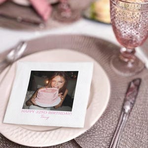 May include: A white square napkin with a printed photo of a person holding a pink birthday cake with a lit candle. The text "HAPPY 22ND BIRTHDAY! Tony" is printed below the photo. The napkin is on a white plate.