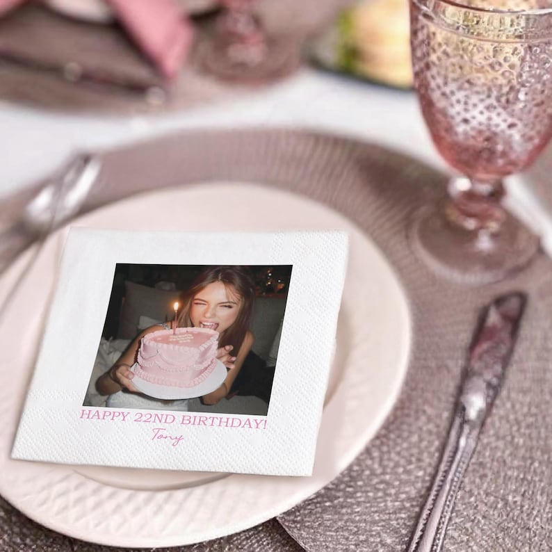 May include: A white square napkin with a printed photo of a person holding a pink birthday cake with a lit candle. The text "HAPPY 22ND BIRTHDAY! Tony" is printed below the photo. The napkin is on a white plate.