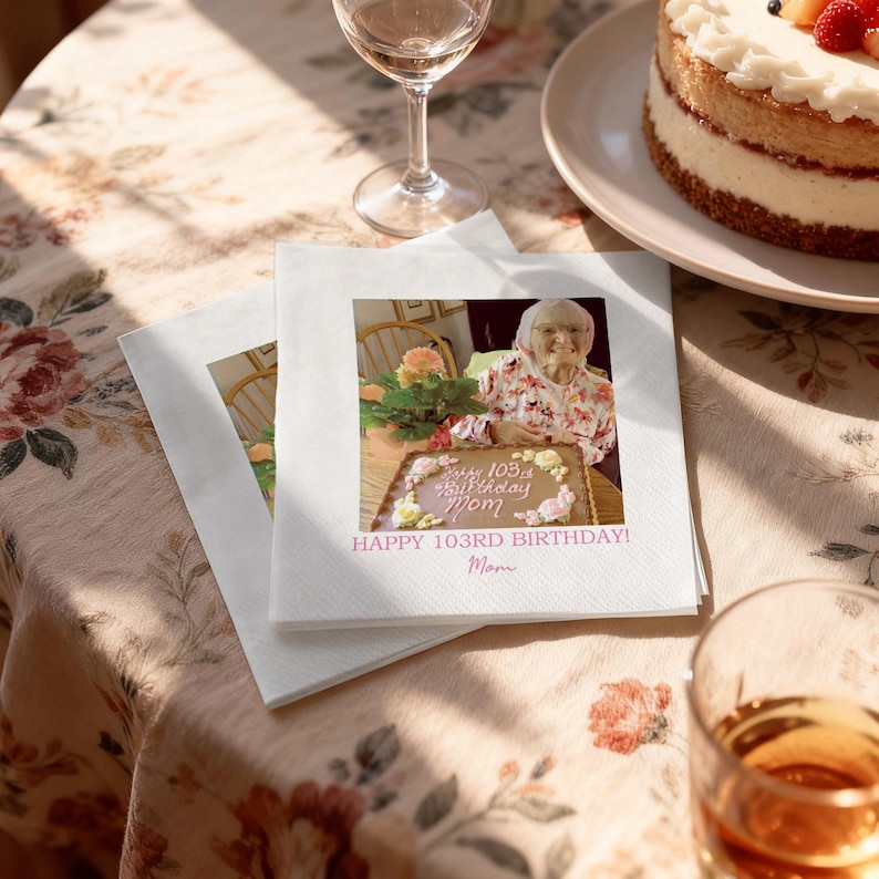 May include: Two white napkins with a printed image of a woman and a cake. The cake has the words "Happy 103rd Birthday Mom" in pink. A partially visible cake and wine glass are in the background.