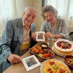 May include: A celebratory table setting with a cake, fried foods, and other dishes. Two people are holding framed photos. The cake is topped with strawberries and a red sauce. The photos have the text "Happy 87th Birthday" and a name.