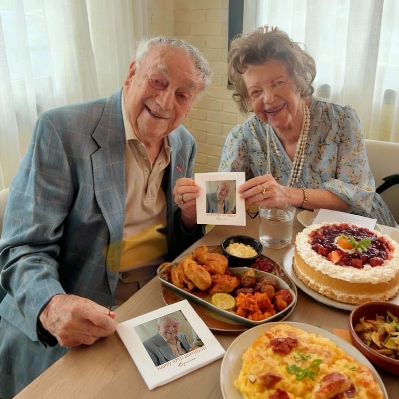 May include: A celebratory table setting with a cake, fried foods, and other dishes. Two people are holding framed photos. The cake is topped with strawberries and a red sauce. The photos have the text "Happy 87th Birthday" and a name.