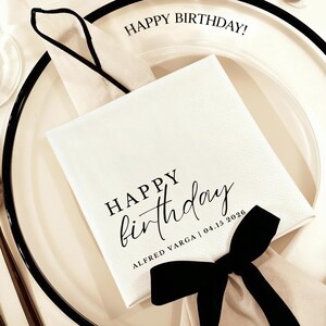 Peut inclure: Une serviette en papier blanche avec l'inscription "HAPPY Birthday" en &eacute;criture noire. La serviette est pos&eacute;e sur une assiette blanche avec un bord noir, et un n&oelig;ud noir est nou&eacute; en bas. Le texte "ALFRED VARGA | 04.15 2026" est &eacute;galement imprim&eacute;.