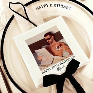 May include: A white square napkin with a photo of a man reading a newspaper and holding a cup of coffee. The napkin has the text "HAPPY BIRTHDAY!" and "HAPPY 30TH BIRTHDAY! Alfred" in black.