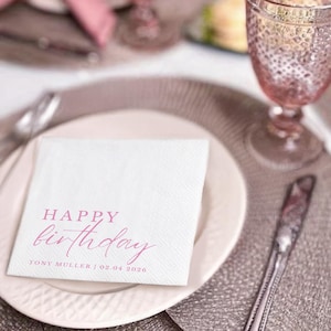 Peut inclure: Une serviette carr&eacute;e blanche avec les mots "HAPPY birthday" imprim&eacute;s en rose. En dessous, on peut lire "TONY MULLER | 02.04.2026". La serviette est pos&eacute;e sur une assiette blanche, avec un verre et des couverts en arri&egrave;re-plan.