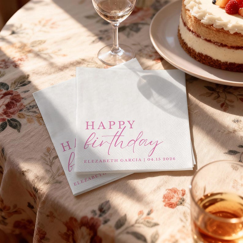 Peut inclure: Deux serviettes en papier blanches avec l'inscription "HAPPY birthday" en rose. Les serviettes sont pos&eacute;es sur une nappe &agrave; motifs floraux, avec un g&acirc;teau et un verre de vin en arri&egrave;re-plan. Le texte "ELEZABETH GARCIA | 04.15 2026" est &eacute;galement imprim&eacute;.