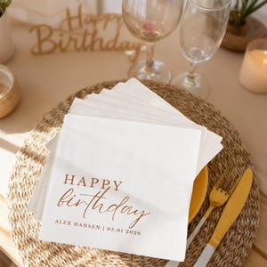 Peut inclure: Serviettes en papier blanches avec l'inscription "Happy Birthday" en marron. Les serviettes sont empil&eacute;es sur un dessous de plat tress&eacute;, avec une fourchette et un couteau dor&eacute;s. Deux verres &agrave; vin sont en arri&egrave;re-plan.