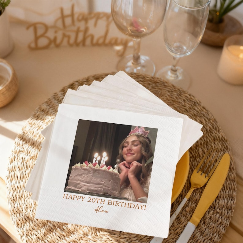 May include: White square napkins with a photo of a person wearing a crown and a birthday cake. The text "HAPPY 20TH BIRTHDAY! Alex" is printed on the napkins. The napkins are on a woven placemat with gold silverware.