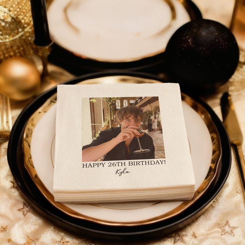 May include: A stack of white paper napkins with a printed photo of a person holding a glass of wine. The text "HAPPY 26TH BIRTHDAY! Kyle" is printed below the photo. The napkins are on a plate with a gold rim.