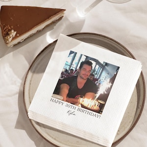 May include: A stack of white paper napkins with a printed photo and text that reads "HAPPY 30TH BIRTHDAY! Kyle." A slice of chocolate cake with a white cream topping sits on a plate.