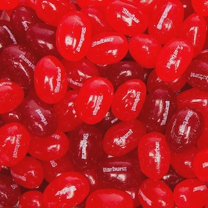 May include: A close-up shot of a pile of red Starburst candies. The candies are oval-shaped and have a glossy appearance. The Starburst logo is visible on many of the candies. The candies are a mix of light and dark red.
