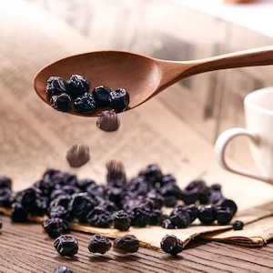 May include: A wooden spoon holding dark blue dried blueberries, with some berries falling. The spoon is over a pile of blueberries on a wooden surface. A white mug is in the background.