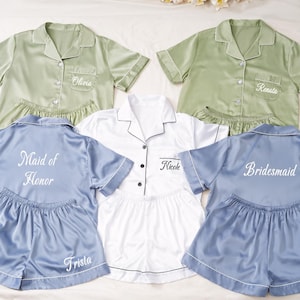 Könnte beinhalten: Passende Pyjama-Sets in Salbeigrün, Weiß und Blau. Jedes Set besteht aus einem kurzärmeligen Hemd mit Knöpfen und Shorts. Einige Hemden sind mit Namen und Titeln wie "Maid of Honor" und "Bridesmaid" bestickt.
