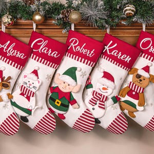 Op de afbeelding: Vijf gepersonaliseerde kerstsokken met de namen Alyssa, Clara, Robert, Karen en Oscar. Elke sok is wit met een rode bovenkant en gestreepte manchet, met een ander feestelijk karakter: rendier, kat, elf, sneeuwman en hond.