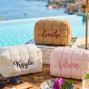 May include: Three quilted cosmetic bags in beige, white, and pink, each embroidered with a name: "Emily", "Kayla", and "Olivia". The bags are on a wooden table with sunglasses, jewelry, and a small dish with flowers.