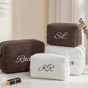 May include: A collection of rectangular cosmetic bags with a waffle-textured fabric. The set includes dark brown and white bags, each featuring custom embroidered initials or names in a cursive script font, secured with a silver-toned zipper closure.