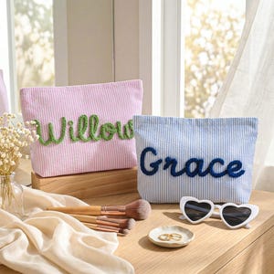 May include: Two personalized seersucker cosmetic bags, one pink with "Willow" in green script and the other blue with "Grace" in blue script. Makeup brushes, heart-shaped sunglasses, and a small dish with rings are also on display.