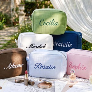 Puede incluir: Seis neceseres personalizados en varios colores, incluyendo verde, blanco, azul marino, marrón, azul claro y rosa. Cada neceser tiene un nombre bordado en un color complementario. Los neceseres están colocados sobre una mesa con maquillaje y perfume.