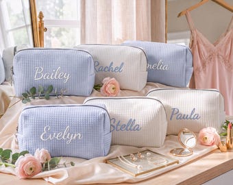 May include: A collection of rectangular waffle-textured cosmetic bags in light blue and white, each featuring embroidered names including Bailey, Rachel, Stella, and Paula. The bags are arranged with pink flowers and jewelry.